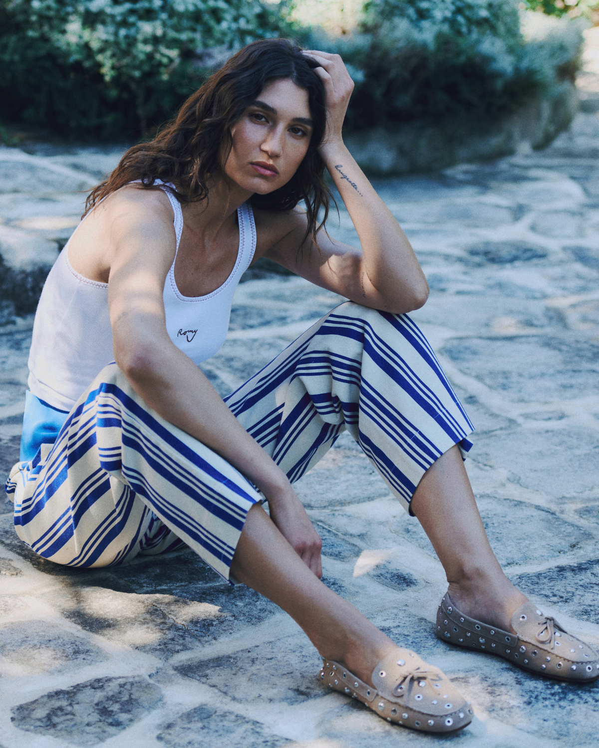 Model seated outdoors wearing ROMY River Pants in blue and cream stripes, styled with a white tank top.