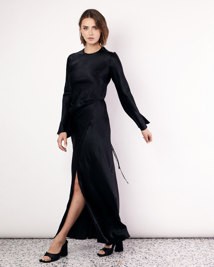 The Long Sleeve Wrap Dress boasts a fluid form and elevated style, featuring gathered detailing and a leg split. It is crafted from a 100% Crushed Acetate in Black. Now available at Romy. 