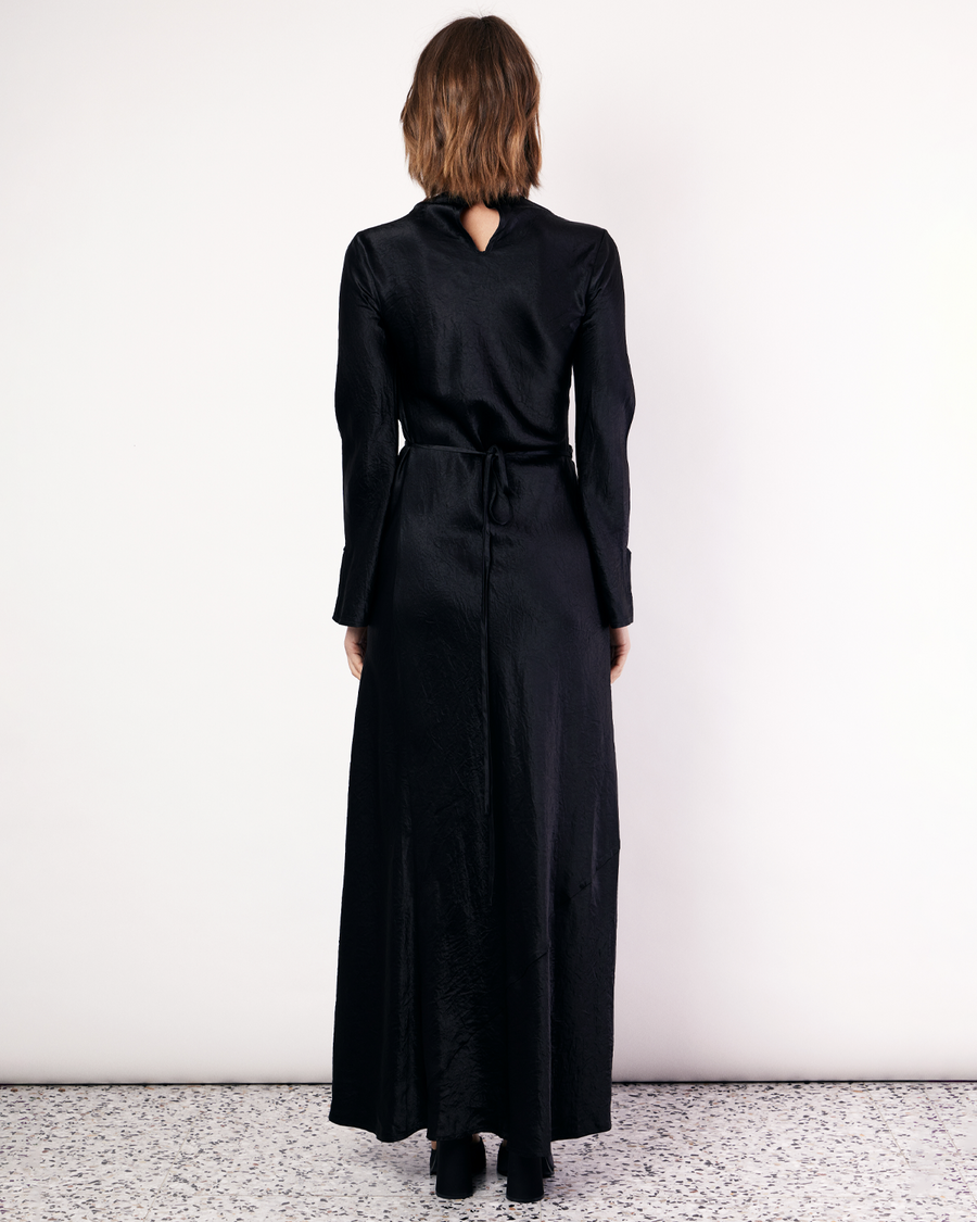 The Long Sleeve Wrap Dress boasts a fluid form and elevated style, featuring gathered detailing and a leg split. It is crafted from a 100% Crushed Acetate in Black. Now available at Romy. 